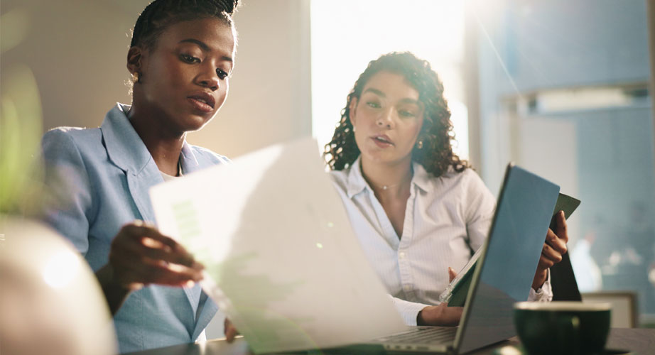 Two finance professionals collaborating around a desk with laptops and documents, discussing data and strategy in a modern office environment.