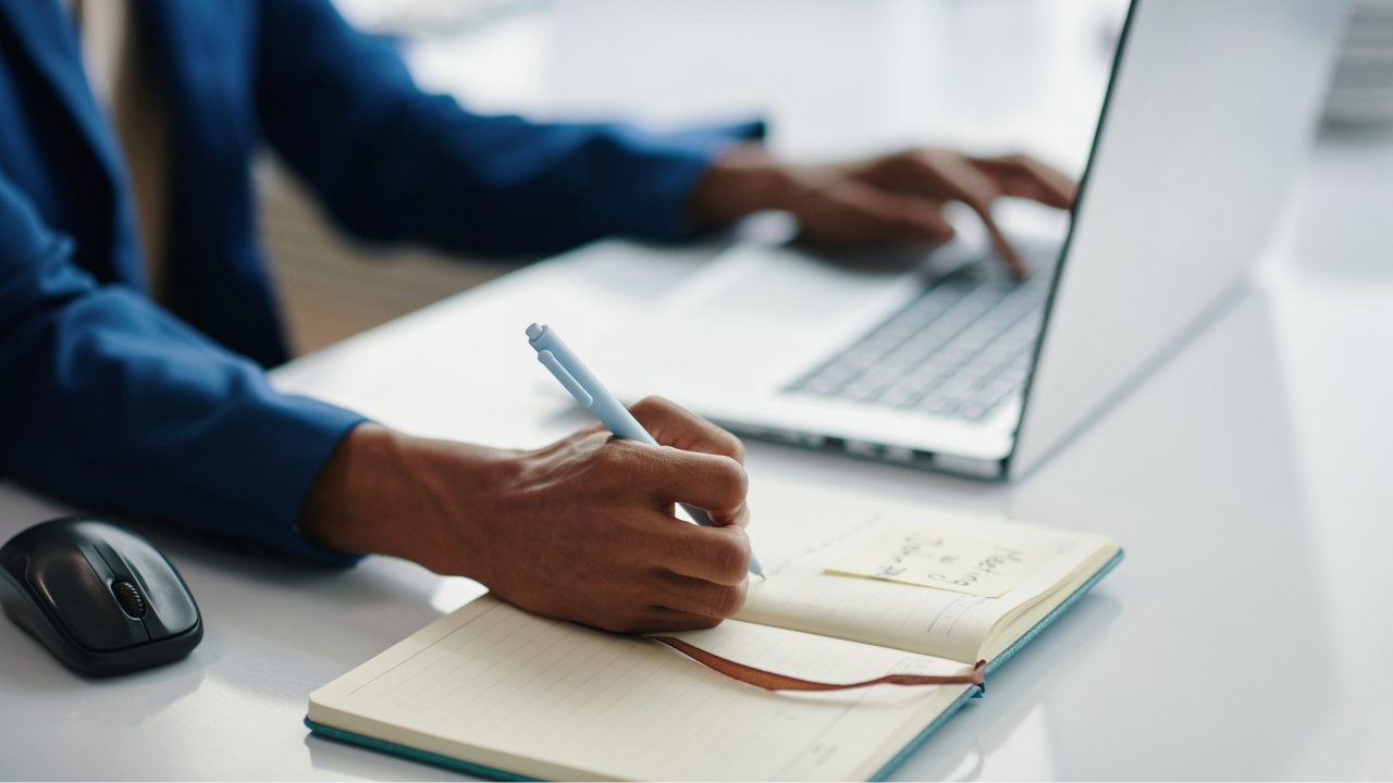 Person taking notes beside a laptop while comparing CRM options