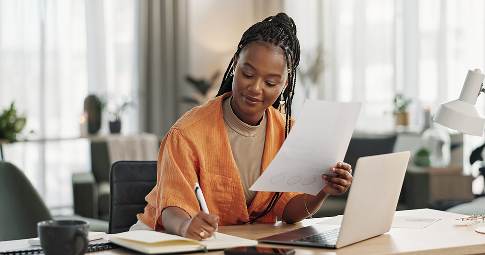 A young woman is taking notes with a pen and notepad. She is wearing an orange cardigan and on the desk is an open laptop.