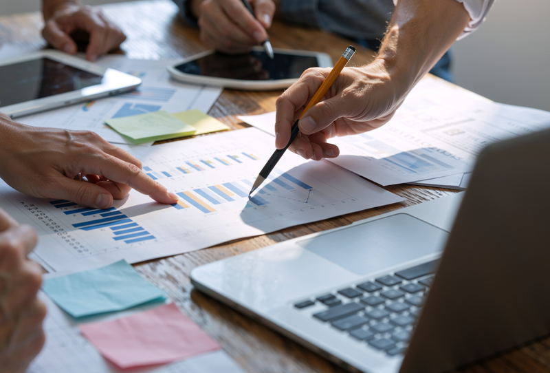 Team members gathered around scattered documents and laptops, illustrating disorganised information and collaboration challenges within an organisation.