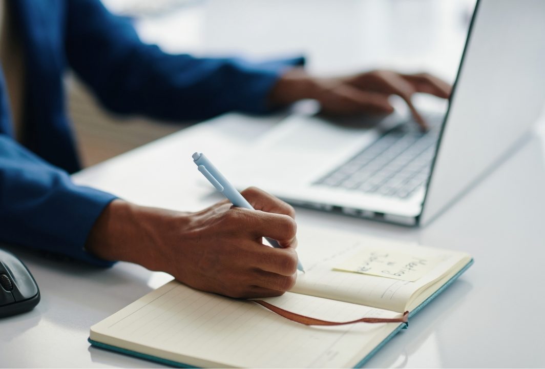 Person taking notes beside a laptop while comparing CRM options